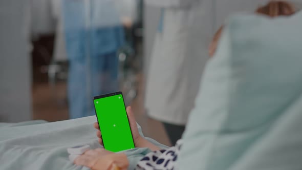 Hospitalized Little Child Resting in Bed Looking at Mock Up Green Screen Chroma Key Smartphone