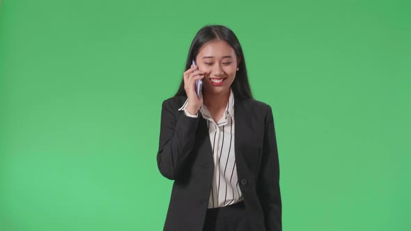 A Smiling Asian Business Woman Talking On Mobile Phone While Walking On Green Screen Studio