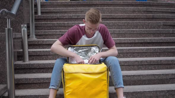 Front View Portrait Handsome Courier Unzipping Yellow Food Delivery Bag Thinking Sitting on Urban