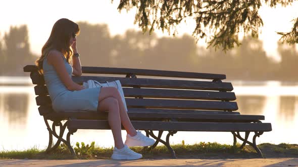 Young Woman in Summer Dress Sitting Relaxed on Lake Side Bench on Warm Evening