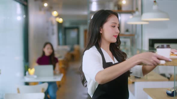 Attractive asian female cafe worker wears apron giving takeaway food bag to customer.