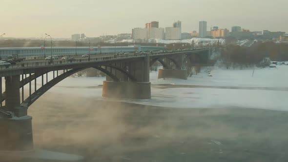 Steam Rise Over River Covered in Ice and Snow. Cars Ride Over Bridge. Nice Day.