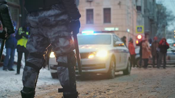 Armored Policeman with Batons Stands in Front of Car with Flashing Strobe Lights