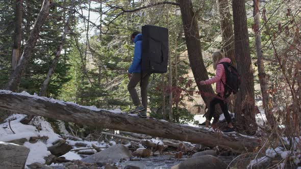 Couple hiking with crash pad as they balance on log crossing a river