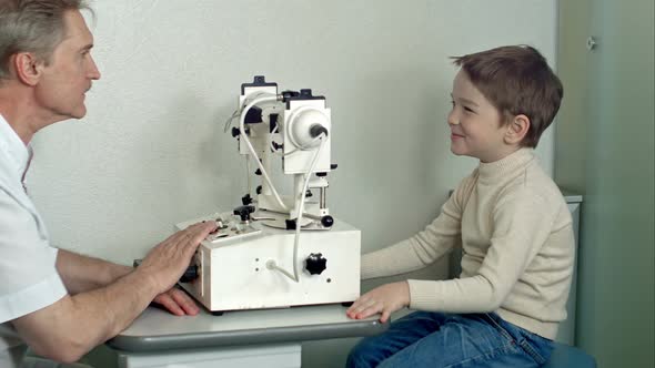 Ophthalmologist in Exam Room with Little Boy Sitting in Chair Looking Into Eye Test Machine