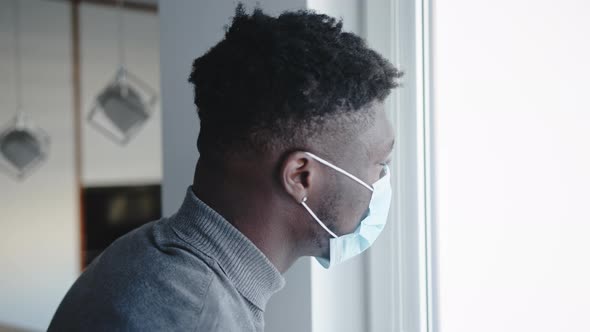 Black Man Wearing Medical Mask Standing By Window Looking Outside. Quarantine and Mental Health