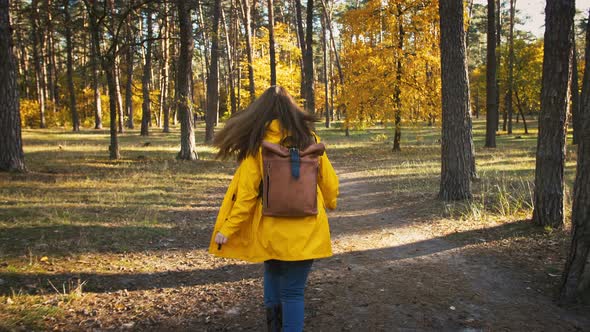 Unknown Young Woman in Parka Coat and Backpack is Running By a Pathway in Autumn Wood on Sunny Day