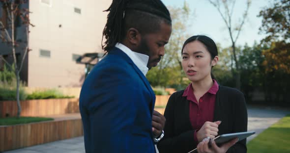 Businessman and Asian Woman Look Into Tablet on Street