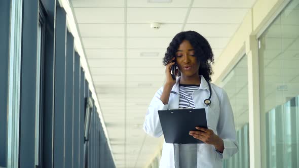 Smiling African American Nurse Talking on the Phone with a Notebook in a Corridor of a Modern