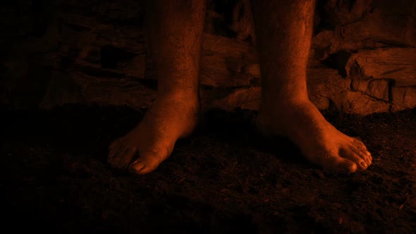 Man Stands Barefoot In Firelight On Dirt Floor