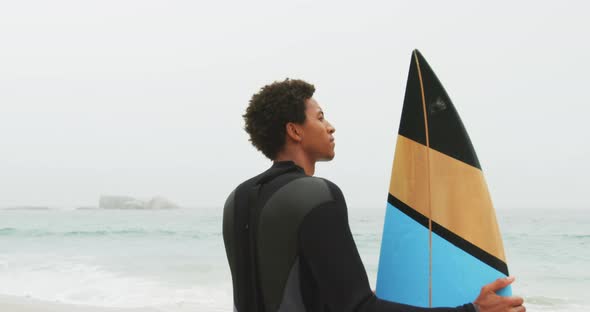 Rear view of African American male surfer standing with surfboard on the beach 4k