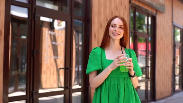 Portrait of Smiling Attractive Young Woman Drinking Cocktail Through Straw Standing at City Street