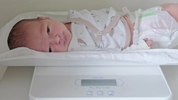 Weighing newborn baby in white clothes on a home scale