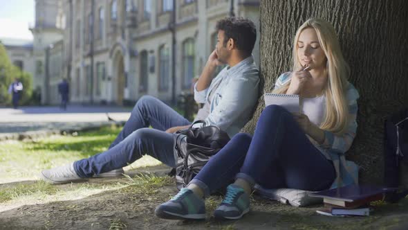 Multiracial Guy Talking on Cellphone Under Tree, Girl Taking Notes in Notebook