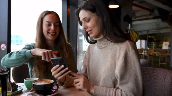 Two Young Beautiful Girl Friends in a Cafe are Looking at the Phone Smiling and Laughing