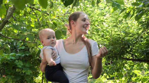 Happy Smiling Mother Holding Her Baby Son and Showing Him Growing Trees in Orchard