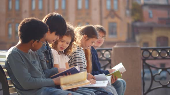 Happy Diverse School Children Reading Books Sitting on Wooden Street Bench Outdoors