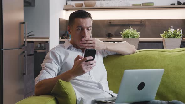 Shocked Man Reacting Bad Message on Smartphone Covers His Mouth in Fright Sitting on Sofa