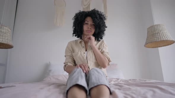 Interested Young African American Woman Sitting on Bed Looking at Camera Listening to Video Chat