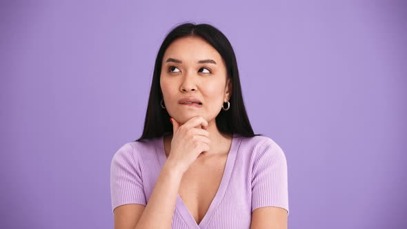 Positive Asian brunette woman wearing purple t-shirt thinking