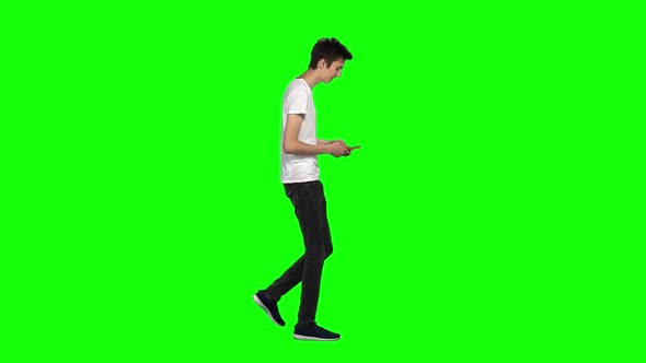 Tall Skinny Teen Guy Calmly Walking and Texting Message Vie His Mobile Phone on Green Screen. Slow