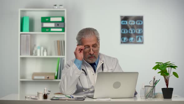 Aged Male Doctor in White Medical Coat Consults Remote Patient Online on Laptop Computer