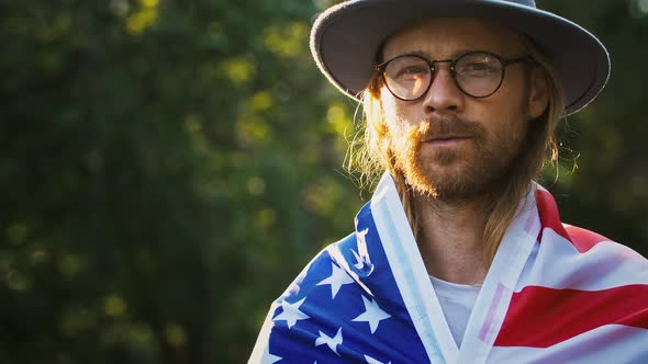 Young Longhaired Fellow in Casual Outfit Glasses and Hat is Wrapping in Flag of USA and Looking at