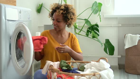 Beautiful Young Woman Putting Clothes Into Washing Machine Sitting on Floor in Laundry Room Spbd