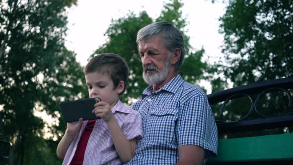 Senior Grandfather and Grandson Are Sitting on a Bench in the Park and Playing on a Smartphone