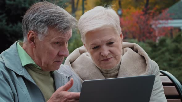 Caucasian Senior Couple Mature Wife with Husband Sit on Bench Outdoors Use Wifi on Laptop Elderly