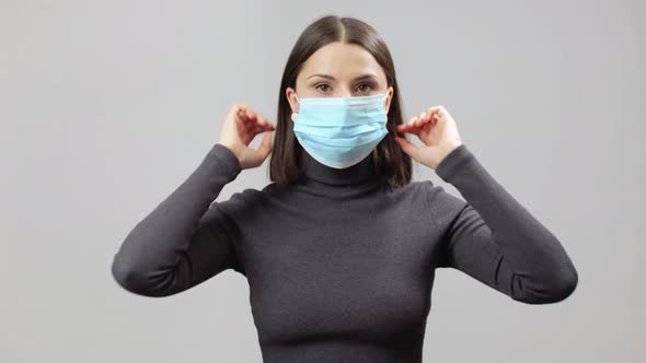 Caucasian Woman In Gray Turtle Neck Dress On A Grey Background Put A Too Big Face Mask On