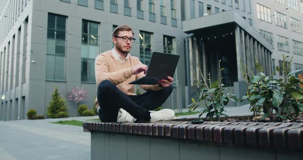 Young Man Using Laptop Sitting Outdoors Near Office Company Building Background.