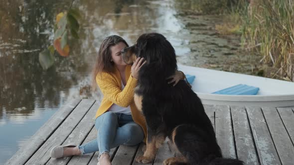 A Young Woman in a Yellow Sweater Is Sitting on a Pier and Stroking a Large Dog