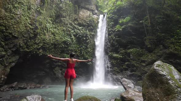 Slim Young Woman in a Red Romper Raising Hands in Front of a Scenic Waterfall.