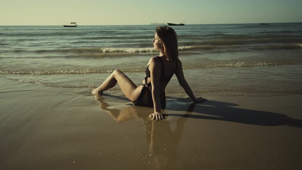 Young Slim Beautiful Redhaired Girl in a White Swimsuit Sunbathes at Sunset Sitting on the Beach