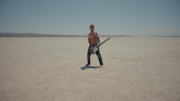 Redhead Barefoot Bloody Musician Plays On Guitar With Chains Around His Neck In Desert