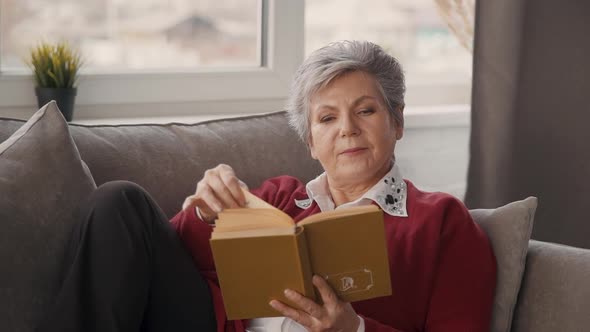 Senior Woman Is Laying on Couch and Reading Interesting Story of True Love and Beautiful Adventure