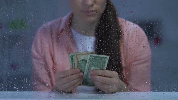 Pensive Poor Middle-Aged Female Counting Dollar Cash and Looking in Rainy Window