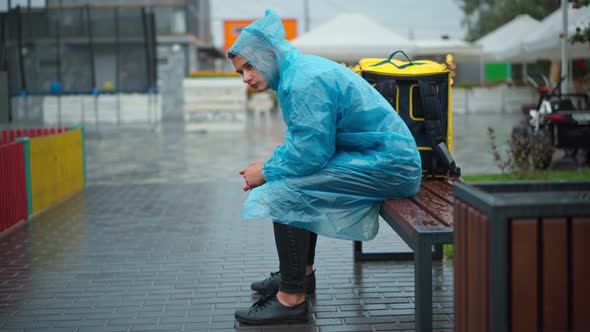 Wide Shot Side View of Sad Young Courier Sitting on Rainy Day in City Waiting