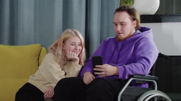 Happy Couple of Blonde Woman and Man in a Wheelchair Looking at the Phone Screen