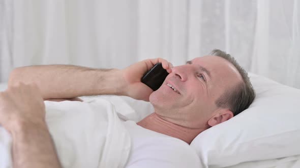 Positive Middle Aged Man Talking on Smartphone in Bed