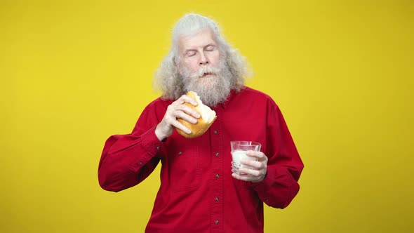 Middle Shot of Senior Greyhaired Bearded Man Eating Bread and Drinking Milk on Yellow Background