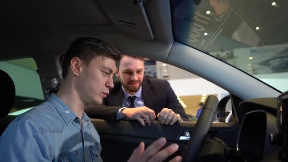 Cheerful Young Man Sitting Behind the Wheel of New Car in Auto Dealership