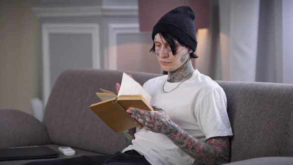 Intelligent Confident Young Tattooed Man Reading Book Sitting at Home on Comfortable Couch in Living