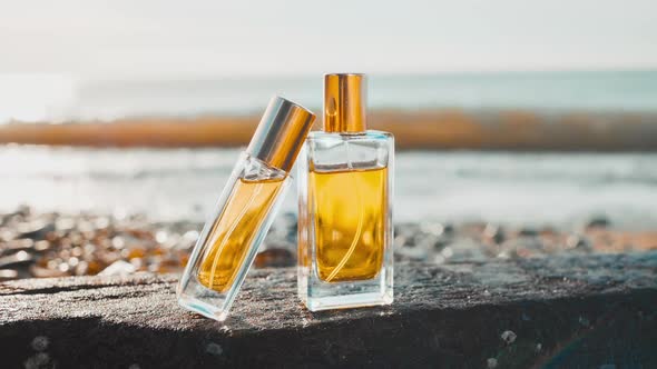 Two glass bottles of golden perfume stand on a rock on the seashore.
