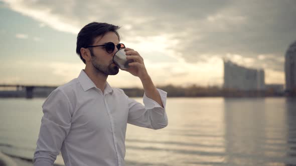 Businessman Drinking Tea Or Coffee From Steel Cup And Enjoys View. Man Relaxing On City Beach.