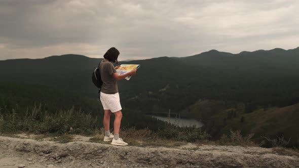 A young girl on a trip stands with a paper map against the background of mountains and a lake.