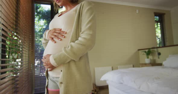 Midsection of caucasian pregnant woman standing at window and touching belly