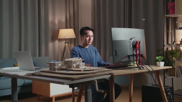 Smiling Asian Male Engineer With The House Model While Working On A Desktop At Home