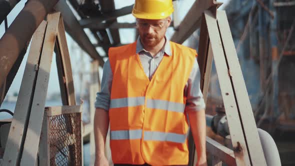 Man shipbuilder in uniform passing over a metal bridge taking off his helmet